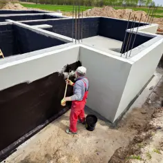 Budowa fundamentów: pracownik maluje ściany ciemną masą izolacyjną.