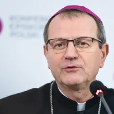 Biskup w okularach i fioletowej zuce, najmłodszy biskup w Polsce, przemawia do mikrofonu.