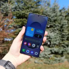 Dłoń trzyma smartfon z interfejsem One UI 8, pokazującym pogodę i powiadomienia na tle drzew.