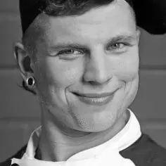 Uśmiechnięty mężczyzna w czapce i białej koszuli, z kolczykiem w uchu. Czy to Ryjek z Warsaw Shore, co się stało?
