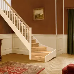 Eleganckie schody w stylu angielskim, z drewnianymi stopniami i jasną balustradą, prowadzą do przytulnego wnętrza z kominkiem i obrazem na ścianie.