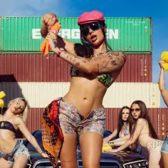 Kobiety w bikini myją samochód wśród kontenerów. W tle widać stosy kontenerów, a na pierwszym planie kilka kobiet w skąpych strojach kąpielowych, trzymających gąbki i spryskiwacze. Jedna z nich, w różowej czapce, trzyma pomarańczową szmatkę. To jakby t...