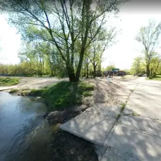Słoneczna plaża nad rzeką, z piaszczystym zejściem do wody, zielenią i drzewami. W oddali widać plac zabaw i niebieskie toalety. To idealne miejsce na relaks, jak na plaży żoliborz.