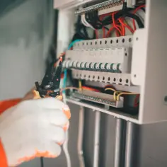 Elektryk w białych rękawicach pracuje przy skrzynce z bezpiecznikami, przygotowując przewody. Widać liczne bezpieczniki i kable, a także narzędzie do obróbki przewodów, które mierzy odpowiednie ampery.