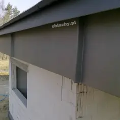 Szara blacha na deskę czołową chroni budynek. Widać fragment okna i białe bloczki.