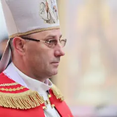 Prymas Polski w uroczystym stroju, w białej mitrze z haftem, w okularach, zamyślony.