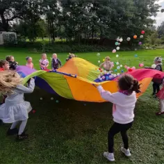 Wesoła animatorka dla dzieci prowadzi zabawę z kolorowym spadochronem i piłeczkami na trawie. Dzieci śmieją się i bawią.