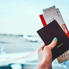 Dłoń trzyma paszport i bilety lotnicze. Na paszporcie widnieje napis "PASSPORT", ale seria i numer są niewidoczne.