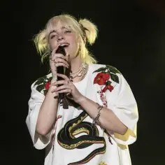 Billie Eilish Polska na koncercie, w białej bluzce z wężem i kwiatami, śpiewa do mikrofonu.
