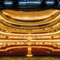 Wielki teatr, plan widowni z rzędami czerwonych foteli, oświetlony ciepłym blaskiem.