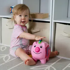 Uśmiechnięte dziecko bawi się różową świnką-skarbonką na tle regału z zabawkami.