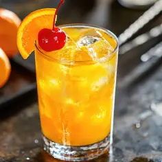 Orzeźwiający drink z pomarańczą i wisienką, podany z lodem. Na tle widać świeże pomarańcze.