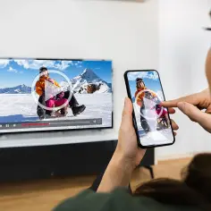 Kobieta korzysta z funkcji screen mirroring, aby wyświetlić film z sankami na dużym ekranie telewizora.