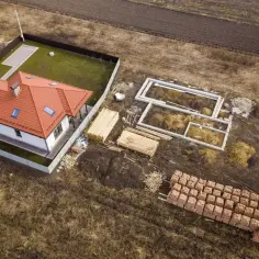 Zabudowa zagrodowa w budowie: gotowy dom z czerwonym dachem obok fundamentów i materiałów budowlanych.
