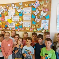 Dzieci z uśmiechem na twarzy pozują przed tablicą pełną kolorowych motyli i cytatów o dobrych uczynkach.