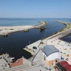 Słoneczny dzień w porcie Kołobrzeg. Statek wypływa na spokojne morze, a plaża tętni życiem.