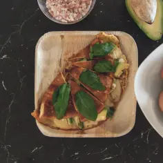 Pyszny omlet z awokado, posypany listkami bazylii, czeka na talerzu. Obok miseczka z jajkami i połówka awokado.
