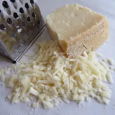 Tarty parmezan w kawałku i świeżo starty ser obok tarki. Wskazówka, jak przechowywać parmezan w kawałku, by zachował świeżość.