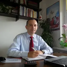 Mężczyzna w jasnej koszuli i czerwonym krawacie siedzi przy biurku, pisząc w notatniku.