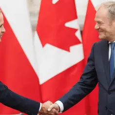 Uśmiechnięci politycy ściskają sobie dłonie na tle flag Kanady. To symboliczne spotkanie, które umacnia więzi między Polską a Kanadą.