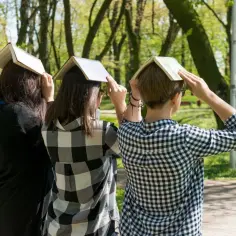 Trzy osoby w parku trzymają książki na głowach, jakby czytały opowiadania Tadeusza Borowskiego.