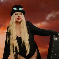 Kobieta w cylindrze i okularach z kolcami, w czarnym stroju i marynarce, z walizką, na tle dramatycznego, pomarańczowego nieba. Niech żyje bal!