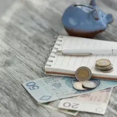 Zapiski o rencie socjalnej z MOPS. Na drewnianym blacie leżą polskie banknoty, monety, notatnik i niebieskowa świnka-skarbonka.