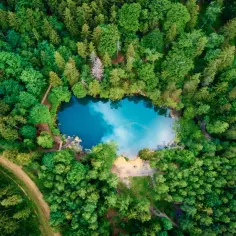 Niebieskie jezioro otoczone gęstym lasem, z piaszczystą plażą i ścieżką prowadzącą do jego brzegów.