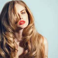 Kobieta z burzą falujących, blond włosów, ułożonych w modny butterfly cut. Jej usta podkreślone są jaskrawą czerwienią.