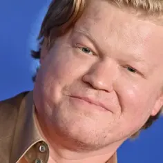 Jesse Plemons, znany z filmów i seriali, w brązowym garniturze na niebieskim tle.