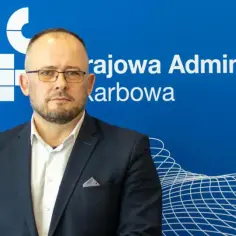 Naczelnik urzędu skarbowego w garniturze, na tle niebieskiej ściany z logo Krajowej Administracji Skarbowej.