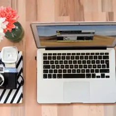 Jak zgrać zdjęcia z aparatu na komputer? Na biurku leży laptop, myszka, aparat i bukiet kwiatów.