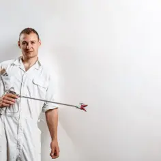 Malarz w kombinezonie trzymający agregat do malowania natryskowego. Dowiedz się, ile kosztuje malowanie natryskowe ścian.
