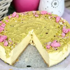 Puszysty sernik w kąpieli wodnej, udekorowany pistacjami i różowymi drażetkami. Idealny na deser.