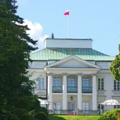 Pałac Belwederski, gdzie mieszka prezydent Polski, otoczony zielenią. Biała fasada z kolumnami i polską flagą na dachu.