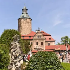 Zamek Czocha, gdzie jest, widać zza kamiennej rzeźby i zieleni. Turyści zwiedzają dziedziniec.
