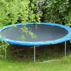 Jak zabezpieczyć trampolinę przed wiatrem? Okrągła trampolina z niebieską osłoną sprężyn stoi na trawie, otoczona zielenią.