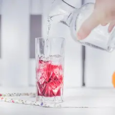 Ręka wlewa wodę z dzbanka do szklanki z lodem i czerwonym napojem. Obok pomarańcza.