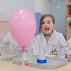 Dzieci w fartuchach laboratoryjnych z ekscytacją obserwują eksperymenty chemiczne. Różowy balon unosi się nad stołem pełnym probówek i płynów.