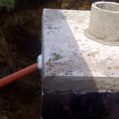 Pomarańczowa rura do szamba wychodzi z wykopu i wchodzi w betonową konstrukcję. Obok betonowy krąg studzienki.