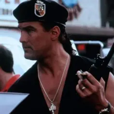 Steven Seagal w beretce i z naszyjnikiem, trzymający krótkofalówkę. Scena z jednego z jego filmów akcji.