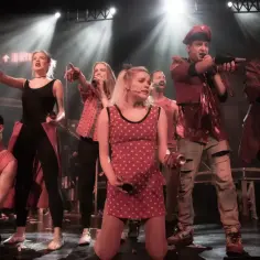 Aktorzy w czerwonych strojach śpiewają na scenie, tworząc energetyczny musical metro.