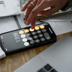 Dłoń z pierścionkiem na palcu wpisuje liczby na kalkulatorze w telefonie, pomagając obliczyć VAT do zapłaty. Obok leży laptop i dokumenty.