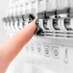Palec naciska na bezpiecznik w skrzynce elektrycznej. W tle widać kolejne zabezpieczenia i symbole gniazdek oraz żarówki. rbk.