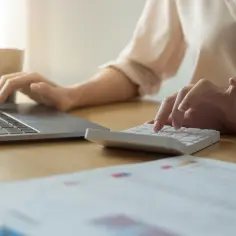 Kobieta liczy na kalkulatorze, sprawdzając, ile kosztuje rzeczoznawca majątkowy. Na biurku laptop i dokumenty.
