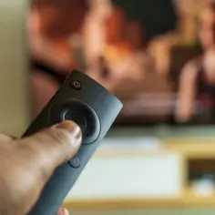 Dłoń trzyma pilot, a na ekranie telewizora widać ludzi. Czy bez anteny można oglądać telewizję? Tak, dzięki platformom streamingowym i dekoderom.