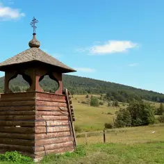 Drewniana dzwonnica z krzyżem na szczycie, otoczona zielonymi wzgórzami. To jedne z wielu regietów atrakcji.