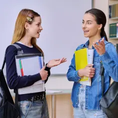 Dwie uczennice rozmawiają o tym, ile jest egzaminów zawodowych w technikum, trzymając książki i plecaki.
