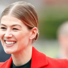 Uśmiechnięta Rosamund Pike, znana z wielu filmów i seriali, w czerwonej marynarce.