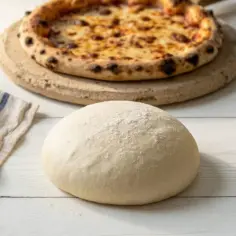 Surowe ciasto na pizzę, gotowa pizza, oliwa z oliwek i ściereczka na białym drewnianym stole.